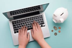Pessoa digitando em um notebook prateado sobre uma superfície azul, com um cofrinho branco e moedas espalhadas ao lado, representando aprendizado financeiro e gestão de dinheiro online.