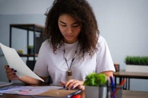Mulher de cabelos cacheados e camiseta branca analisa gráficos e documentos coloridos em uma mesa de escritório. O ambiente tem decoração minimalista, com plantas e materiais de papelaria, transmitindo foco e dedicação em um trabalho analítico ou estudo financeiro.
